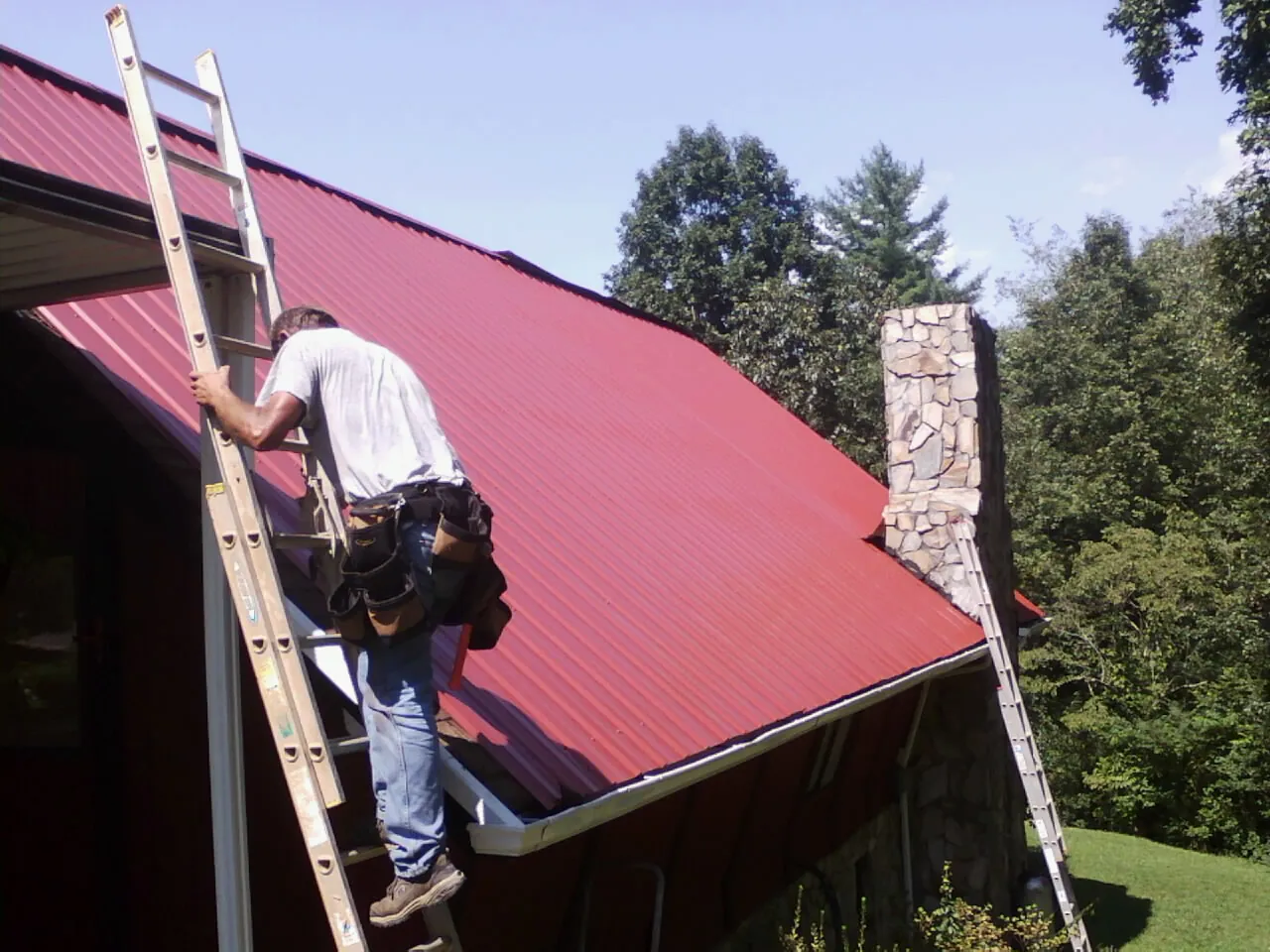 Roofer on a ladder beside a red metal roof and stone chimney — Soffit & Fascia Repair by Roofing Brothers in Angola